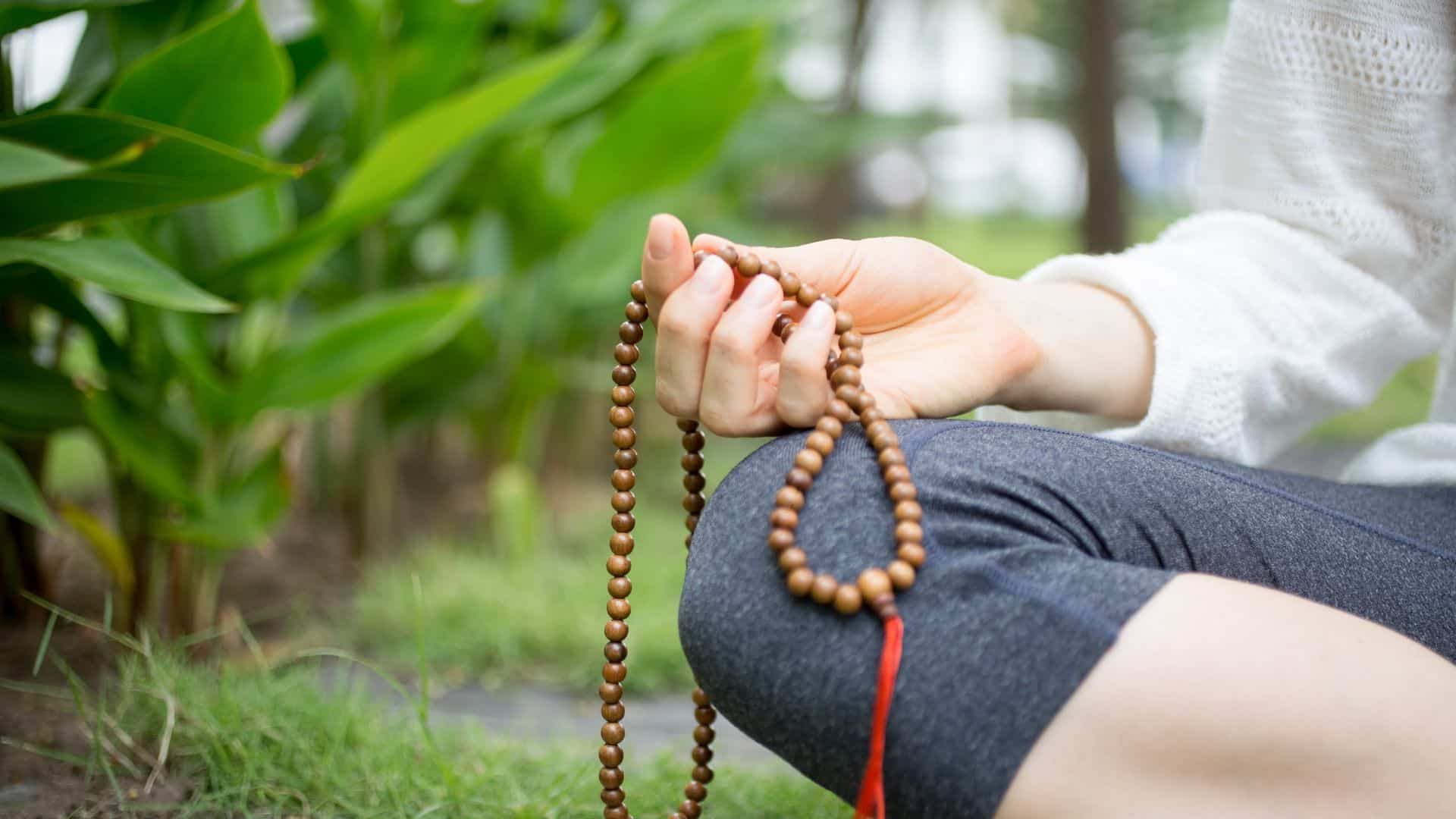A close-up of a person seated in meditation, gently holding mala beads in their hand in a peaceful garden setting—symbolizing the grounding use of a mantra for anxiety.