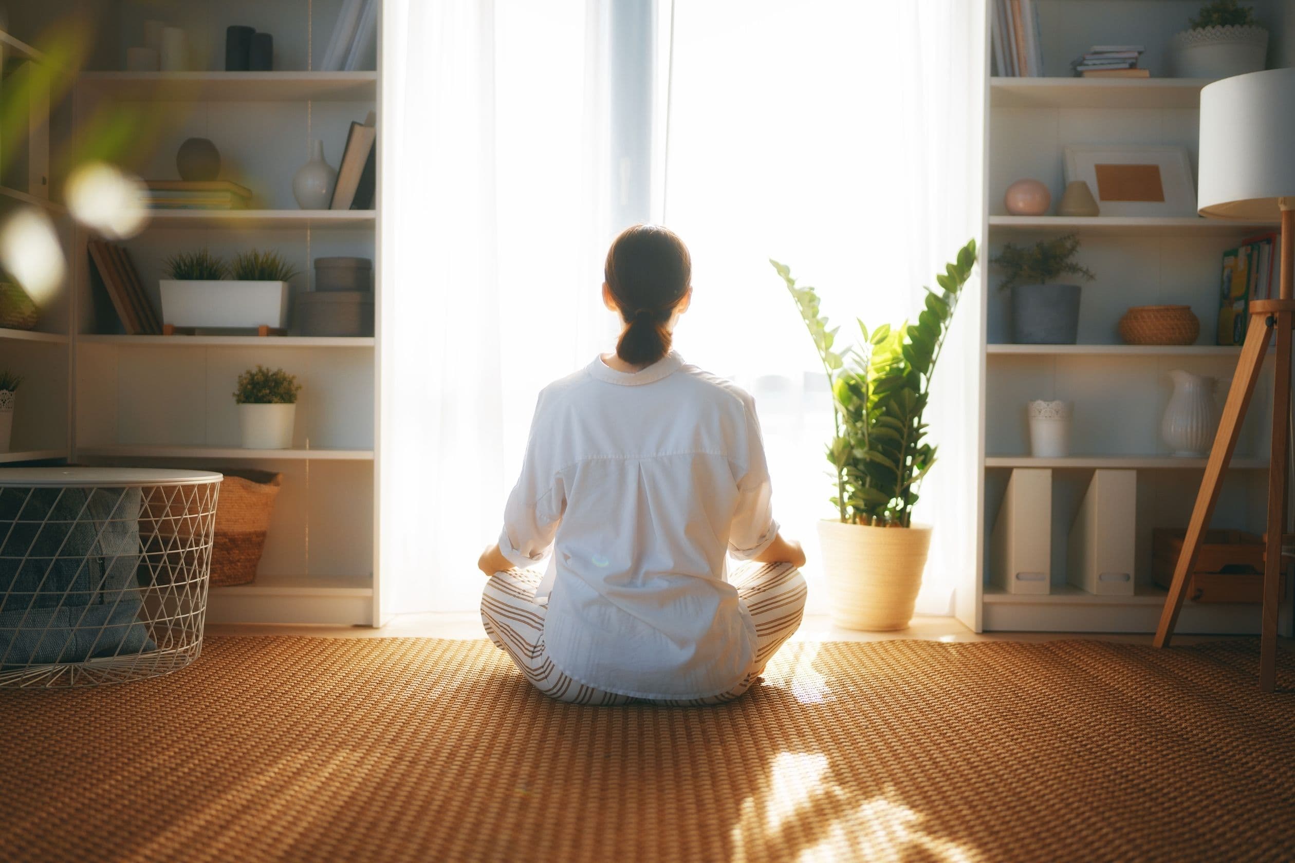 A woman in striped pajamas sits cross-legged on a woven mat, meditating in front of a window with sheer curtains and surrounded by houseplants and soft natural light.