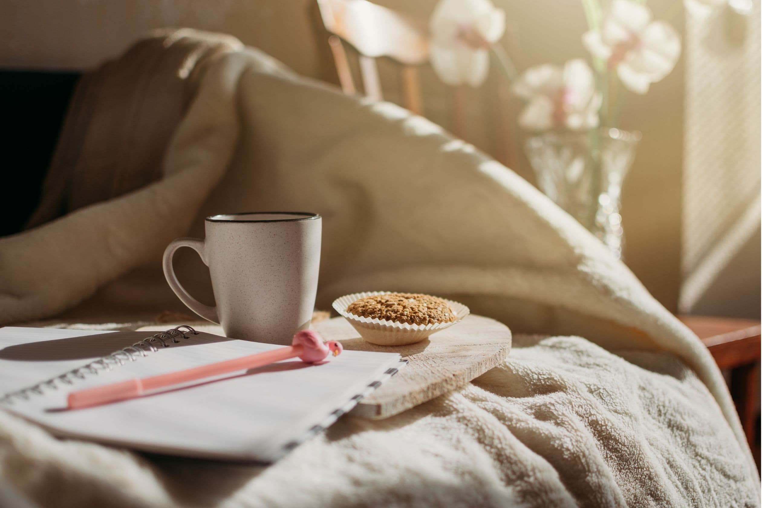 A cozy scene with a warm blanket, journal, pink pen, ceramic mug, and a muffin on a wooden tray, illuminated by soft morning light and orchid flowers in the background.